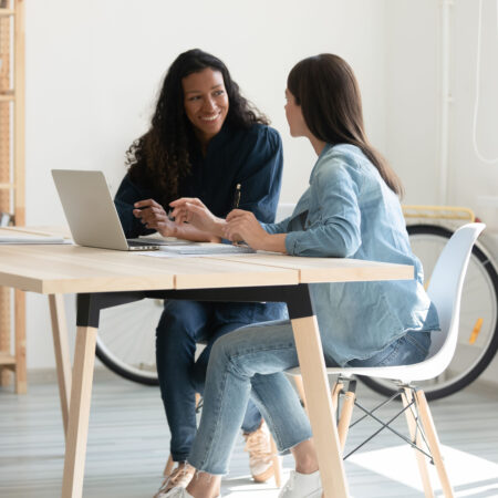 Smiling female colleagues sit at desk in office talk brainstorm work together on laptop