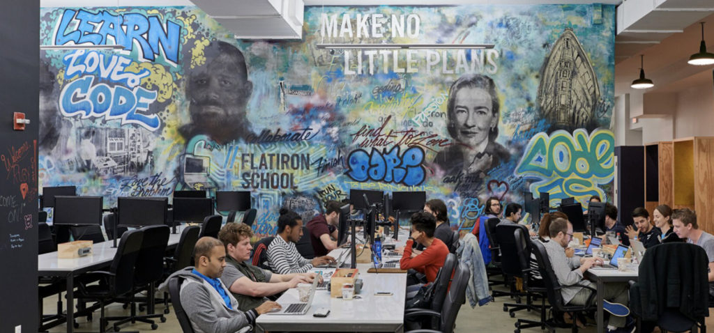 Students sit at two rows of desks with laptops and monitors. A large wall graffitied with various company values provides a colorful backdrop.