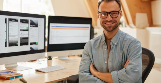 A man smiling at a desk with two computer screens behind him.