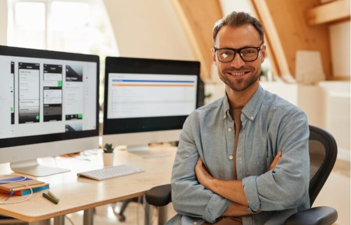 A man smiling at a desk with two computer screens behind him.