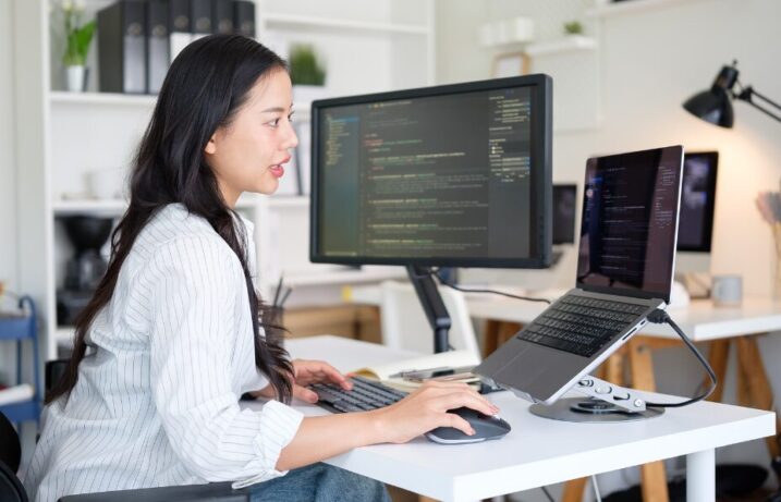A female working at a desk with a computer monitor and laptop displaying code in a modern office setting.